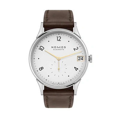 Nomos Minimatik mit weissen Zifferblatt, schwarzen arabischen Zahlen & Minutenpunkte. Die Zeiger sind in goldig Gehalten. Das Datum bei 3 Uhr mit goligem Rahmen, sowie  die kleinen Sekunde, die bei 6 Uhr liegt. Dunkelbraun Lederband rundet das Design ab.