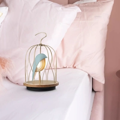 Ein Jingoos auf einem Bett, umgeben von verschiedenen rosa Kissen. Die Wand ist ebenfalls in einem zarten Rosaton gehalten. Der K&auml;fig des Vogels ist goldfarben, und die Keramik im K&auml;fig leuchtet in einem sanften Hellblau.