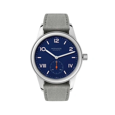 Zentrale Aufnahme der Uhr von vorn. Tiefblaues Zifferblatt mit wei&szlig;en und rosa Ziffern. Kleine rote Sekunde bei 6 Uhr. Zeiger silberfarben. Band aus grauem Wildleder, Geh&auml;use aus silbernem Metall. Marke: &bdquo;NOMOS Glash&uuml;tte&ldquo;. Modern, klar, sachlich.